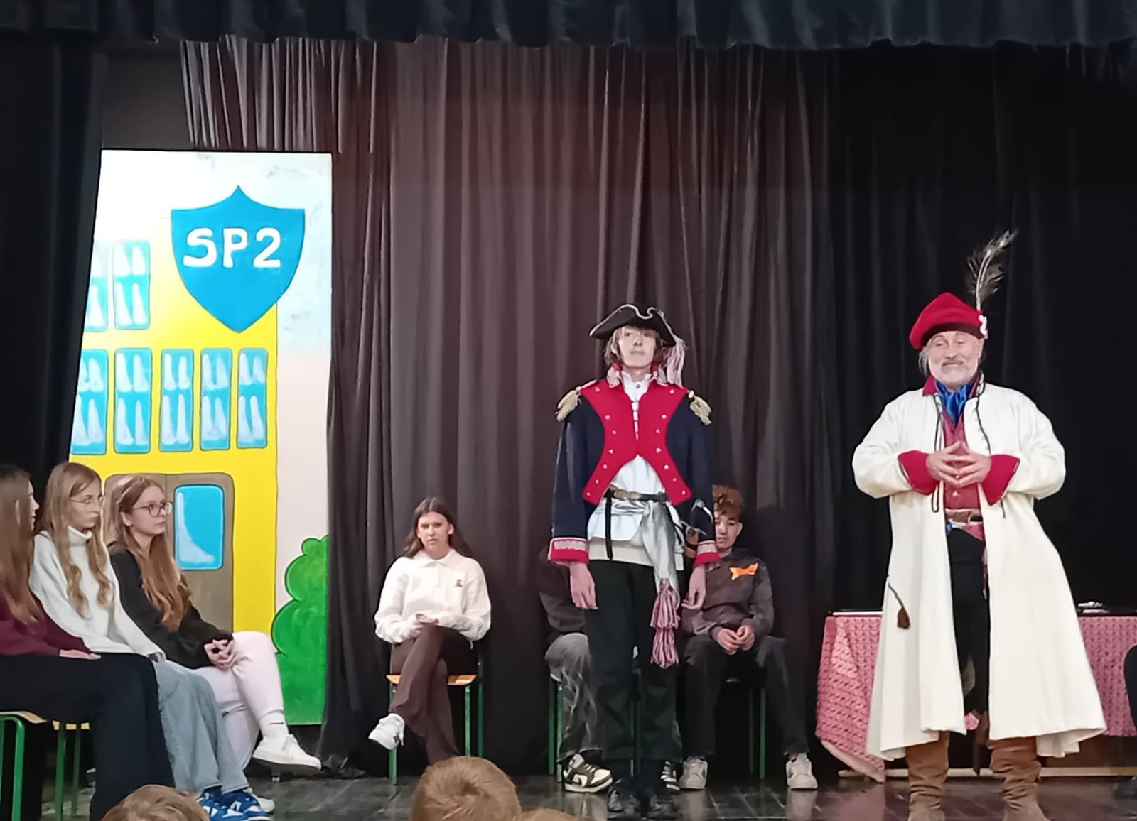 Każdego roku uczniowie naszej szkoły mają niepowtarzalną okazję wziąć udział w wyjątkowym wydarzeniu edukacyjnym - ŻYWEJ LEKCJI HISTORII, prowadzonej przez grupę rekonstrukcyjną Chorągiew Komturstwa Gniewskiego. To spotkanie, które na długo pozostaje w pamięci uczestników, łącząc wiedzę historyczną z emocjami, ruchem i bezpośrednim kontaktem z przeszłością.
W tym roku tematyka spotkań została zrealizowana na dwóch poziomach wiekowych:
Klasy 4-6: Świat starożytnych Greków i Rzymian
Uczniowie klas młodszych przenieśli się do czasów starożytności - epoki potężnych cywilizacji, mitologii, igrzysk olimpijskich i legionów rzymskich. Dzięki strojom z epoki, realistycznym rekwizytom i barwnym opowieściom rekonstruktorów, dzieci mogły poczuć atmosferę antycznego świata. Dowiedzieli się, jak wyglądało życie codzienne Greków i Rzymian, jakie były zwyczaje wojenne, a nawet jak wyglądały ćwiczenia wojskowe legionistów. Zajęcia - prowadzone w formie interaktywnej - dały uczniom możliwość przymierzenia elementów uzbrojenia oraz wzięcia udziału w krótkich pokazach.
Klasy 7-8: Długi wiek XIX
Starsze klasy uczestniczyły w lekcji poświęconej Długiemu wiekowi XIX, czyli okresowi, który ukształtował współczesny świat. Rekonstruktorzy przybliżyli wydarzenia związane z rewolucjami, walką o niepodległość, przemianami społecznymi i narodzinami nowoczesnych idei. Szczególną uwagę poświęcono wydarzeniom ważnym dla Polski - rozbiorom, powstaniom narodowym i postaciom kluczowym dla naszej historii. Uczniowie mogli zobaczyć repliki broni, a także stroje z XIX wieku. Dla wielu była to okazja do poznania historii w sposób bardziej angażujący niż podręcznik - przez opowieści, prezentacje multimedialne i inscenizacje.
Żywa lekcja historii to nie tylko atrakcja, ale przede wszystkim cenna forma nauki. Bezpośredni kontakt z pasjonatami historii oraz aktywne uczestnictwo w przedsięwzięciu sprawia, że uczniowie lepiej zapamiętują materiał, rozbudzają swoją ciekawość i rozwijają szacunek do dziedzictwa kulturowego.