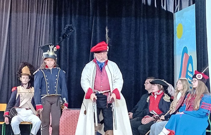 Każdego roku uczniowie naszej szkoły mają niepowtarzalną okazję wziąć udział w wyjątkowym wydarzeniu edukacyjnym - ŻYWEJ LEKCJI HISTORII, prowadzonej przez grupę rekonstrukcyjną Chorągiew Komturstwa Gniewskiego. To spotkanie, które na długo pozostaje w pamięci uczestników, łącząc wiedzę historyczną z emocjami, ruchem i bezpośrednim kontaktem z przeszłością.
W tym roku tematyka spotkań została zrealizowana na dwóch poziomach wiekowych:
Klasy 4-6: Świat starożytnych Greków i Rzymian
Uczniowie klas młodszych przenieśli się do czasów starożytności - epoki potężnych cywilizacji, mitologii, igrzysk olimpijskich i legionów rzymskich. Dzięki strojom z epoki, realistycznym rekwizytom i barwnym opowieściom rekonstruktorów, dzieci mogły poczuć atmosferę antycznego świata. Dowiedzieli się, jak wyglądało życie codzienne Greków i Rzymian, jakie były zwyczaje wojenne, a nawet jak wyglądały ćwiczenia wojskowe legionistów. Zajęcia - prowadzone w formie interaktywnej - dały uczniom możliwość przymierzenia elementów uzbrojenia oraz wzięcia udziału w krótkich pokazach.
Klasy 7-8: Długi wiek XIX
Starsze klasy uczestniczyły w lekcji poświęconej Długiemu wiekowi XIX, czyli okresowi, który ukształtował współczesny świat. Rekonstruktorzy przybliżyli wydarzenia związane z rewolucjami, walką o niepodległość, przemianami społecznymi i narodzinami nowoczesnych idei. Szczególną uwagę poświęcono wydarzeniom ważnym dla Polski - rozbiorom, powstaniom narodowym i postaciom kluczowym dla naszej historii. Uczniowie mogli zobaczyć repliki broni, a także stroje z XIX wieku. Dla wielu była to okazja do poznania historii w sposób bardziej angażujący niż podręcznik - przez opowieści, prezentacje multimedialne i inscenizacje.
Żywa lekcja historii to nie tylko atrakcja, ale przede wszystkim cenna forma nauki. Bezpośredni kontakt z pasjonatami historii oraz aktywne uczestnictwo w przedsięwzięciu sprawia, że uczniowie lepiej zapamiętują materiał, rozbudzają swoją ciekawość i rozwijają szacunek do dziedzictwa kulturowego.