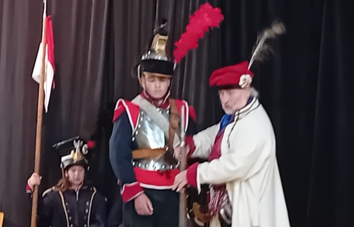 Każdego roku uczniowie naszej szkoły mają niepowtarzalną okazję wziąć udział w wyjątkowym wydarzeniu edukacyjnym - ŻYWEJ LEKCJI HISTORII, prowadzonej przez grupę rekonstrukcyjną Chorągiew Komturstwa Gniewskiego. To spotkanie, które na długo pozostaje w pamięci uczestników, łącząc wiedzę historyczną z emocjami, ruchem i bezpośrednim kontaktem z przeszłością.
W tym roku tematyka spotkań została zrealizowana na dwóch poziomach wiekowych:
Klasy 4-6: Świat starożytnych Greków i Rzymian
Uczniowie klas młodszych przenieśli się do czasów starożytności - epoki potężnych cywilizacji, mitologii, igrzysk olimpijskich i legionów rzymskich. Dzięki strojom z epoki, realistycznym rekwizytom i barwnym opowieściom rekonstruktorów, dzieci mogły poczuć atmosferę antycznego świata. Dowiedzieli się, jak wyglądało życie codzienne Greków i Rzymian, jakie były zwyczaje wojenne, a nawet jak wyglądały ćwiczenia wojskowe legionistów. Zajęcia - prowadzone w formie interaktywnej - dały uczniom możliwość przymierzenia elementów uzbrojenia oraz wzięcia udziału w krótkich pokazach.
Klasy 7-8: Długi wiek XIX
Starsze klasy uczestniczyły w lekcji poświęconej Długiemu wiekowi XIX, czyli okresowi, który ukształtował współczesny świat. Rekonstruktorzy przybliżyli wydarzenia związane z rewolucjami, walką o niepodległość, przemianami społecznymi i narodzinami nowoczesnych idei. Szczególną uwagę poświęcono wydarzeniom ważnym dla Polski - rozbiorom, powstaniom narodowym i postaciom kluczowym dla naszej historii. Uczniowie mogli zobaczyć repliki broni, a także stroje z XIX wieku. Dla wielu była to okazja do poznania historii w sposób bardziej angażujący niż podręcznik - przez opowieści, prezentacje multimedialne i inscenizacje.
Żywa lekcja historii to nie tylko atrakcja, ale przede wszystkim cenna forma nauki. Bezpośredni kontakt z pasjonatami historii oraz aktywne uczestnictwo w przedsięwzięciu sprawia, że uczniowie lepiej zapamiętują materiał, rozbudzają swoją ciekawość i rozwijają szacunek do dziedzictwa kulturowego.