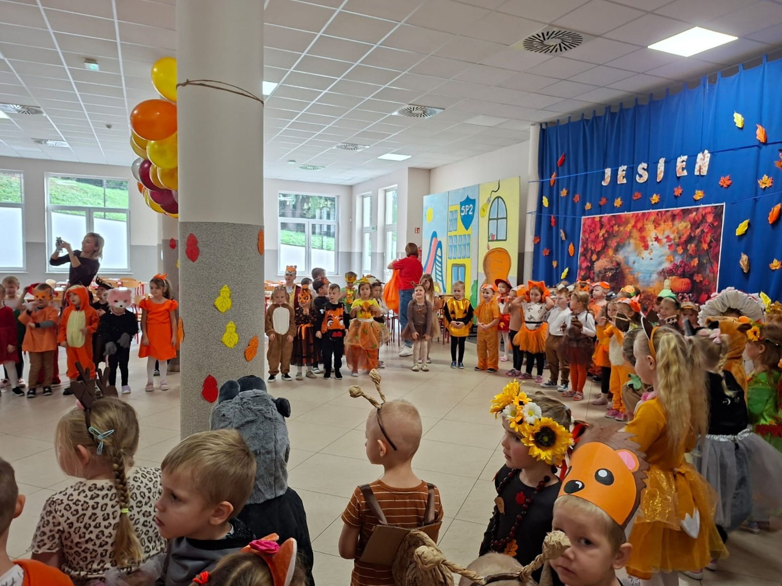 W dniu dzisiejszym w naszym przedszkolu odbył się wyjątkowy i długo wyczekiwany Bal Jesieni. W wydarzeniu wzięły udział wszystkie grupy: Biedronki, Sówki, Leśne Skrzaty oraz Pszczółk oraz dzieci z grup Krasnele i Sówki z Przedszkola Samorządowego nr 1 w Pasłęku, które u nas goszczą od września.
Od samego rana w przedszkolu panowała radosna atmosfera – dzieci przybyły w przepięknych, kolorowych strojach inspirowanych jesienią. Wśród nich można było zobaczyć dynie, jeże, ślimaki, liście, kasztanki, grzybki, wiewiórki, a nawet same Pani Jesienie!
Po powitaniu przez Panie Dyrektor i Wicedyrektor, rozpoczęły się jesienne zabawy i tańce. Wszystkie dzieci wykazały się ogromnym zaangażowaniem, a cała uroczystość odbywała się w duchu radości i wspólnej zabawy.
Nie zabrakło również piosenek i tańców o jesieni – rozbrzmiały takie przeboje z repertuaru dziecięcego Wspólne śpiewanie i tańce przyniosły wszystkim mnóstwo uśmiechu i energii.
Ponadto odbyła się pamiątkowa sesja zdjęciowa w jesiennej scenerii – wśród kolorowych liści, koszy z darami jesieni i szerokich uśmiechów dzieci. 
Jednym z najważniejszych punktów dnia był pokaz jesiennych strojów na niebieskim dywanie. Dzieci z dumą prezentowały swoje przebrania, pozując do zdjęć niczym prawdziwe gwiazdy jesiennego pokazu mody. Publiczność nagradzała ich gromkimi brawami, a w powietrzu czuć było radość i dumę z własnych kreacji.
Bal Jesieni był dniem pełnym barw, muzyki, śmiechu i wspólnej zabawy. Takie wydarzenia integrują dzieci, uczą współpracy, rozwijają kreatywność i pozwalają dostrzec piękno otaczającej nas przyrody. 