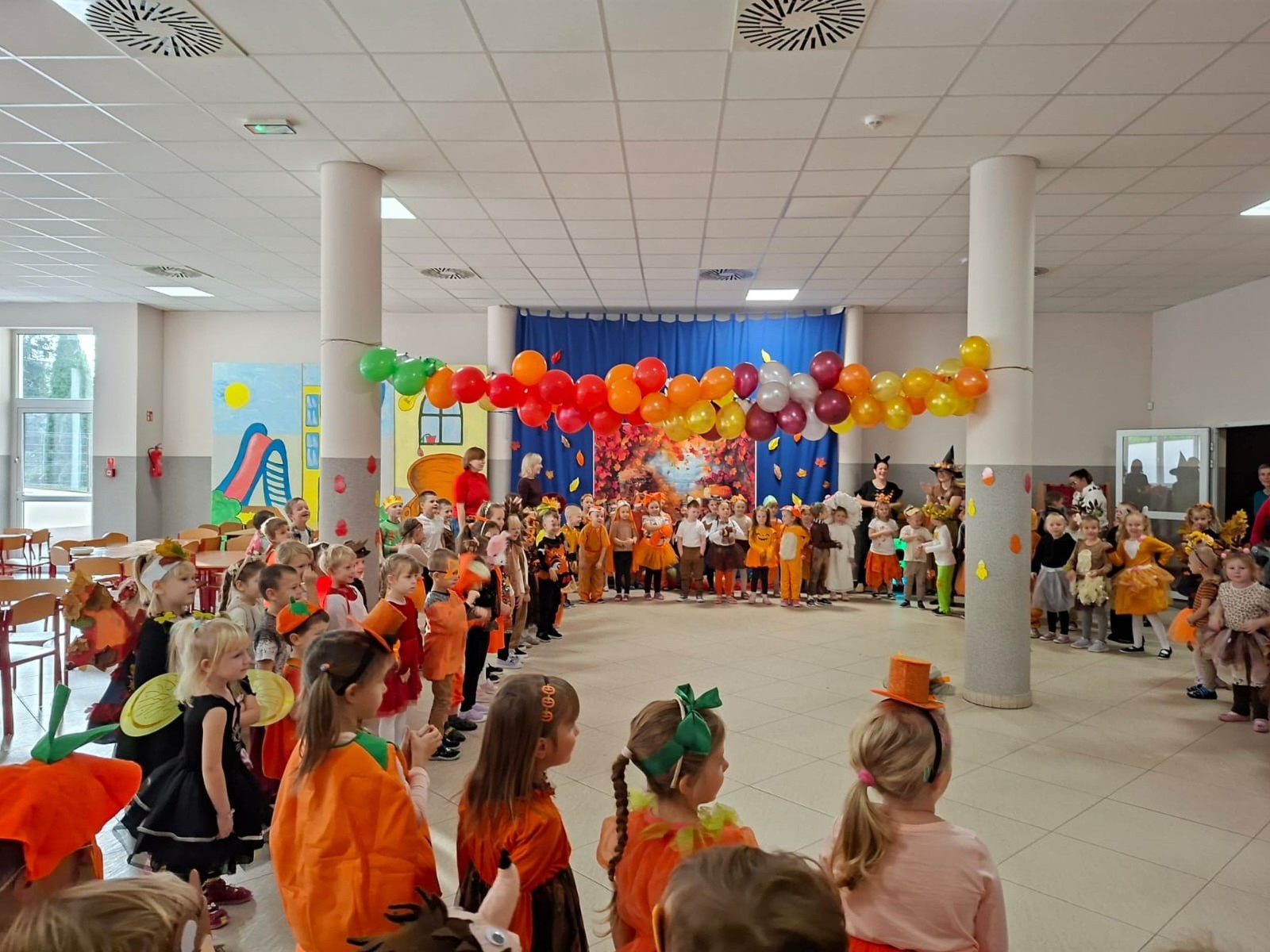 W dniu dzisiejszym w naszym przedszkolu odbył się wyjątkowy i długo wyczekiwany Bal Jesieni. W wydarzeniu wzięły udział wszystkie grupy: Biedronki, Sówki, Leśne Skrzaty oraz Pszczółk oraz dzieci z grup Krasnele i Sówki z Przedszkola Samorządowego nr 1 w Pasłęku, które u nas goszczą od września.
Od samego rana w przedszkolu panowała radosna atmosfera – dzieci przybyły w przepięknych, kolorowych strojach inspirowanych jesienią. Wśród nich można było zobaczyć dynie, jeże, ślimaki, liście, kasztanki, grzybki, wiewiórki, a nawet same Pani Jesienie!
Po powitaniu przez Panie Dyrektor i Wicedyrektor, rozpoczęły się jesienne zabawy i tańce. Wszystkie dzieci wykazały się ogromnym zaangażowaniem, a cała uroczystość odbywała się w duchu radości i wspólnej zabawy.
Nie zabrakło również piosenek i tańców o jesieni – rozbrzmiały takie przeboje z repertuaru dziecięcego Wspólne śpiewanie i tańce przyniosły wszystkim mnóstwo uśmiechu i energii.
Ponadto odbyła się pamiątkowa sesja zdjęciowa w jesiennej scenerii – wśród kolorowych liści, koszy z darami jesieni i szerokich uśmiechów dzieci. 
Jednym z najważniejszych punktów dnia był pokaz jesiennych strojów na niebieskim dywanie. Dzieci z dumą prezentowały swoje przebrania, pozując do zdjęć niczym prawdziwe gwiazdy jesiennego pokazu mody. Publiczność nagradzała ich gromkimi brawami, a w powietrzu czuć było radość i dumę z własnych kreacji.
Bal Jesieni był dniem pełnym barw, muzyki, śmiechu i wspólnej zabawy. Takie wydarzenia integrują dzieci, uczą współpracy, rozwijają kreatywność i pozwalają dostrzec piękno otaczającej nas przyrody. 