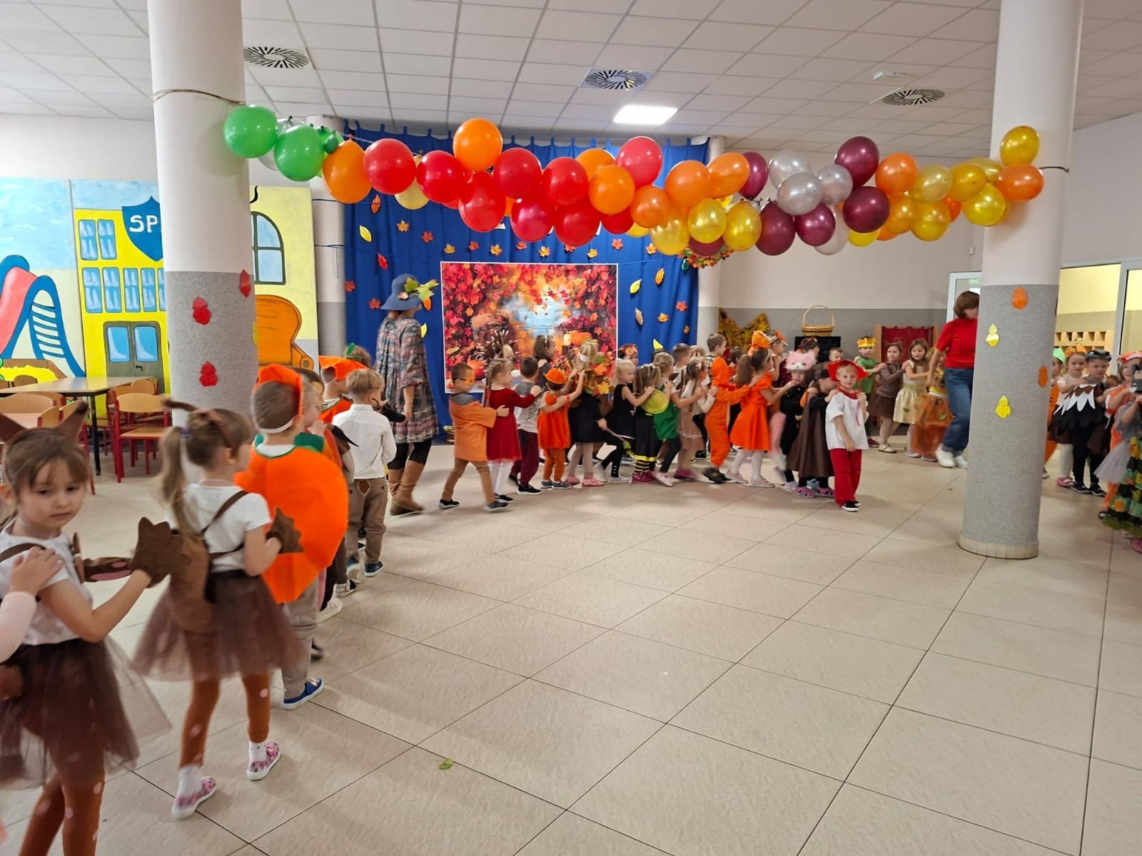 W dniu dzisiejszym w naszym przedszkolu odbył się wyjątkowy i długo wyczekiwany Bal Jesieni. W wydarzeniu wzięły udział wszystkie grupy: Biedronki, Sówki, Leśne Skrzaty oraz Pszczółk oraz dzieci z grup Krasnele i Sówki z Przedszkola Samorządowego nr 1 w Pasłęku, które u nas goszczą od września.
Od samego rana w przedszkolu panowała radosna atmosfera – dzieci przybyły w przepięknych, kolorowych strojach inspirowanych jesienią. Wśród nich można było zobaczyć dynie, jeże, ślimaki, liście, kasztanki, grzybki, wiewiórki, a nawet same Pani Jesienie!
Po powitaniu przez Panie Dyrektor i Wicedyrektor, rozpoczęły się jesienne zabawy i tańce. Wszystkie dzieci wykazały się ogromnym zaangażowaniem, a cała uroczystość odbywała się w duchu radości i wspólnej zabawy.
Nie zabrakło również piosenek i tańców o jesieni – rozbrzmiały takie przeboje z repertuaru dziecięcego Wspólne śpiewanie i tańce przyniosły wszystkim mnóstwo uśmiechu i energii.
Ponadto odbyła się pamiątkowa sesja zdjęciowa w jesiennej scenerii – wśród kolorowych liści, koszy z darami jesieni i szerokich uśmiechów dzieci. 
Jednym z najważniejszych punktów dnia był pokaz jesiennych strojów na niebieskim dywanie. Dzieci z dumą prezentowały swoje przebrania, pozując do zdjęć niczym prawdziwe gwiazdy jesiennego pokazu mody. Publiczność nagradzała ich gromkimi brawami, a w powietrzu czuć było radość i dumę z własnych kreacji.
Bal Jesieni był dniem pełnym barw, muzyki, śmiechu i wspólnej zabawy. Takie wydarzenia integrują dzieci, uczą współpracy, rozwijają kreatywność i pozwalają dostrzec piękno otaczającej nas przyrody. 