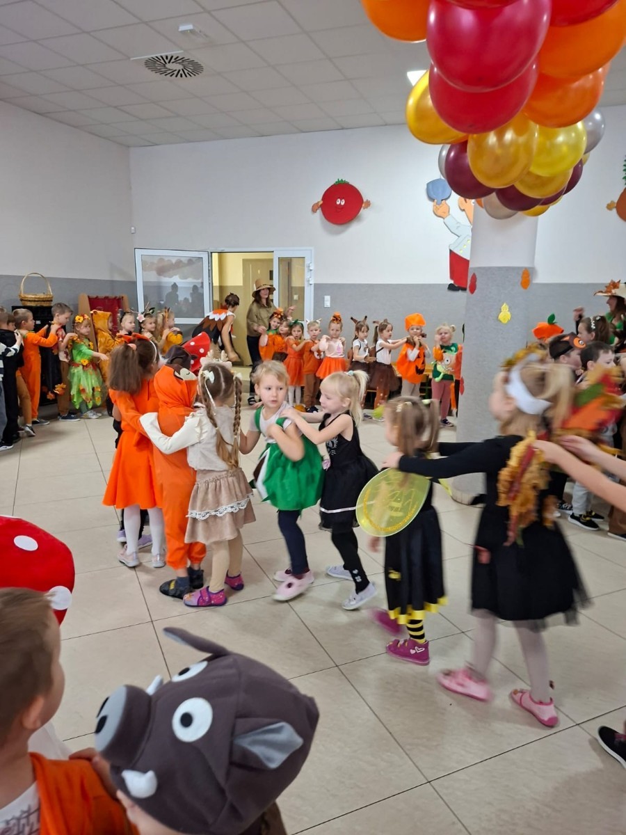 W dniu dzisiejszym w naszym przedszkolu odbył się wyjątkowy i długo wyczekiwany Bal Jesieni. W wydarzeniu wzięły udział wszystkie grupy: Biedronki, Sówki, Leśne Skrzaty oraz Pszczółk oraz dzieci z grup Krasnele i Sówki z Przedszkola Samorządowego nr 1 w Pasłęku, które u nas goszczą od września.
Od samego rana w przedszkolu panowała radosna atmosfera – dzieci przybyły w przepięknych, kolorowych strojach inspirowanych jesienią. Wśród nich można było zobaczyć dynie, jeże, ślimaki, liście, kasztanki, grzybki, wiewiórki, a nawet same Pani Jesienie!
Po powitaniu przez Panie Dyrektor i Wicedyrektor, rozpoczęły się jesienne zabawy i tańce. Wszystkie dzieci wykazały się ogromnym zaangażowaniem, a cała uroczystość odbywała się w duchu radości i wspólnej zabawy.
Nie zabrakło również piosenek i tańców o jesieni – rozbrzmiały takie przeboje z repertuaru dziecięcego Wspólne śpiewanie i tańce przyniosły wszystkim mnóstwo uśmiechu i energii.
Ponadto odbyła się pamiątkowa sesja zdjęciowa w jesiennej scenerii – wśród kolorowych liści, koszy z darami jesieni i szerokich uśmiechów dzieci. 
Jednym z najważniejszych punktów dnia był pokaz jesiennych strojów na niebieskim dywanie. Dzieci z dumą prezentowały swoje przebrania, pozując do zdjęć niczym prawdziwe gwiazdy jesiennego pokazu mody. Publiczność nagradzała ich gromkimi brawami, a w powietrzu czuć było radość i dumę z własnych kreacji.
Bal Jesieni był dniem pełnym barw, muzyki, śmiechu i wspólnej zabawy. Takie wydarzenia integrują dzieci, uczą współpracy, rozwijają kreatywność i pozwalają dostrzec piękno otaczającej nas przyrody. 
