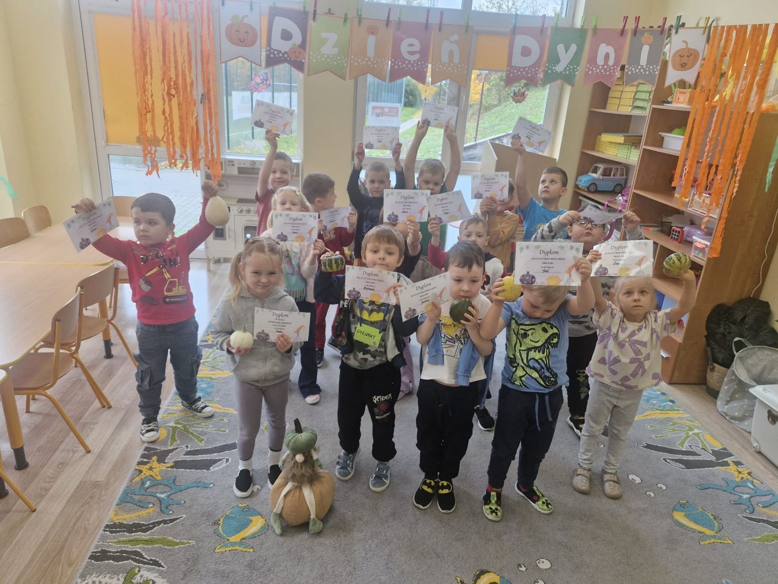 31 października nasze przedszkole zamieniło się w prawdziwą dyniową krainę! W salach królował kolor pomarańczowy, a wszędzie można było zobaczyć uśmiechnięte buzie dzieci i  dynie w różnych kształtach i rozmiarach.Przedszkolaki chętnie brały udział w zabawach muzyczno-ruchowych oraz zajęciach plastycznych z dynią w roli głównej.
Dzieci dowiedziały się, że dynia to nie tylko piękna ozdoba jesieni, ale również zdrowe i smaczne warzywo, z którego można przygotować wiele pysznych potraw.Na zakończenie dnia dzieci otrzymały dyniowe medale oraz pamiątkowe dyplomy,świadczące o ich udziale i zaangażowaniu w obchody tego święta.
To był dzień pełen radości, kolorów ,niezapomnianych wrażeń i jesiennej magii.
