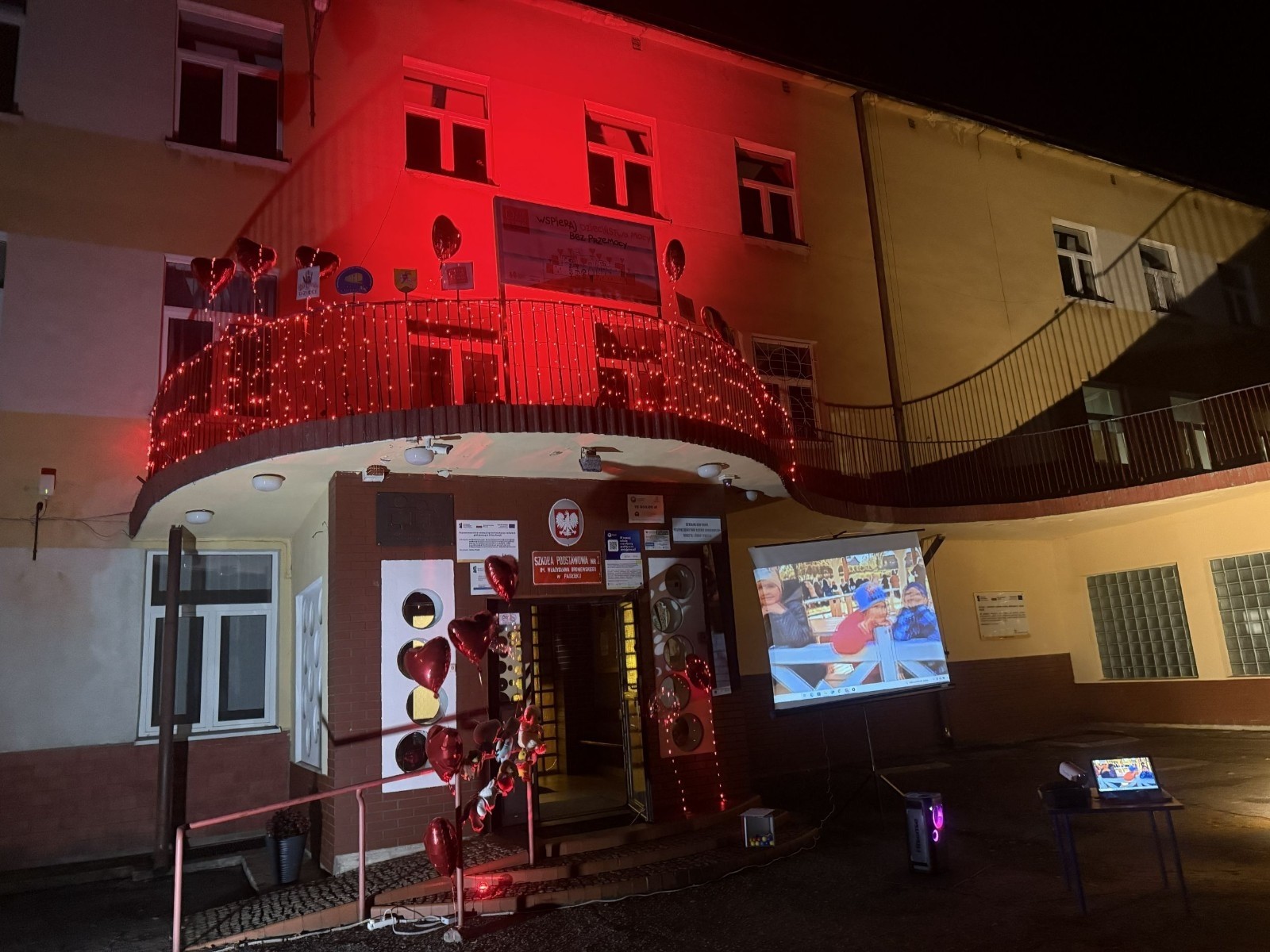 O godzinie 17:00 pod szkołą rozpoczęła się impreza, która zgromadziła około 200 osób. Szkoła została podświetlona na czerwono, co symbolizowało naszą jedność w walce z przemocą. Był to akcent podsumowujący nasze wcześ iej sze  działania w ramach Kampanii Ogólnopolskiej „Dzieciństwo bez Przemocy”. Od października uczniowie uczestniczyli w lekcjach online przygotowanych przez Fundację Dajemy Dzieciom Siłę. Rodzice również zostali objęci wsparciem w postaci webinaru online ww. fudacji.
Uczestnicy mogli wziąć udział w wspólnym śpiewaniu, quizie wiedzy o przemocy oraz tworzeniu „kamieni mocy” – symboli pozytywnej energii i wsparcia. Dodatkowo, w ramach konkursów, przyznano nagrody ufundowane przez Urząd Miejski w Pasłęku.
Ten cudowny czas umilił nam smaczny poczęstunek, w tym grochówka, pizza i ciasta, gorąca kawa i herbatka przygotowany przez panie kucharki i rodziców.
W wydarzeniu uczestniczyli również zaproszeni goście, w tym Burmistrz Pasłęka – pani Elżbieta Wasiuk,  podinspektor KP Pasłęk – pani Agnieszka Bielewicz - Piechowiak oraz przedstawiciel Kuratorium Oświaty w Elblągu – pani Teresa Bednarz, którzy wspierają nasze działania. Ich obecność była dowodem na to, jak ważna jest współpraca lokalnej społeczności w walce z przemocą wobec dzieci. Dziękujemy wszystkim, którzy wzięli udział w tym wyjątkowym dniu oraz przyczynili się do jego organizacji. Razem możemy stworzyć bezpieczne i przyjazne środowisko dla naszych dzieci!
