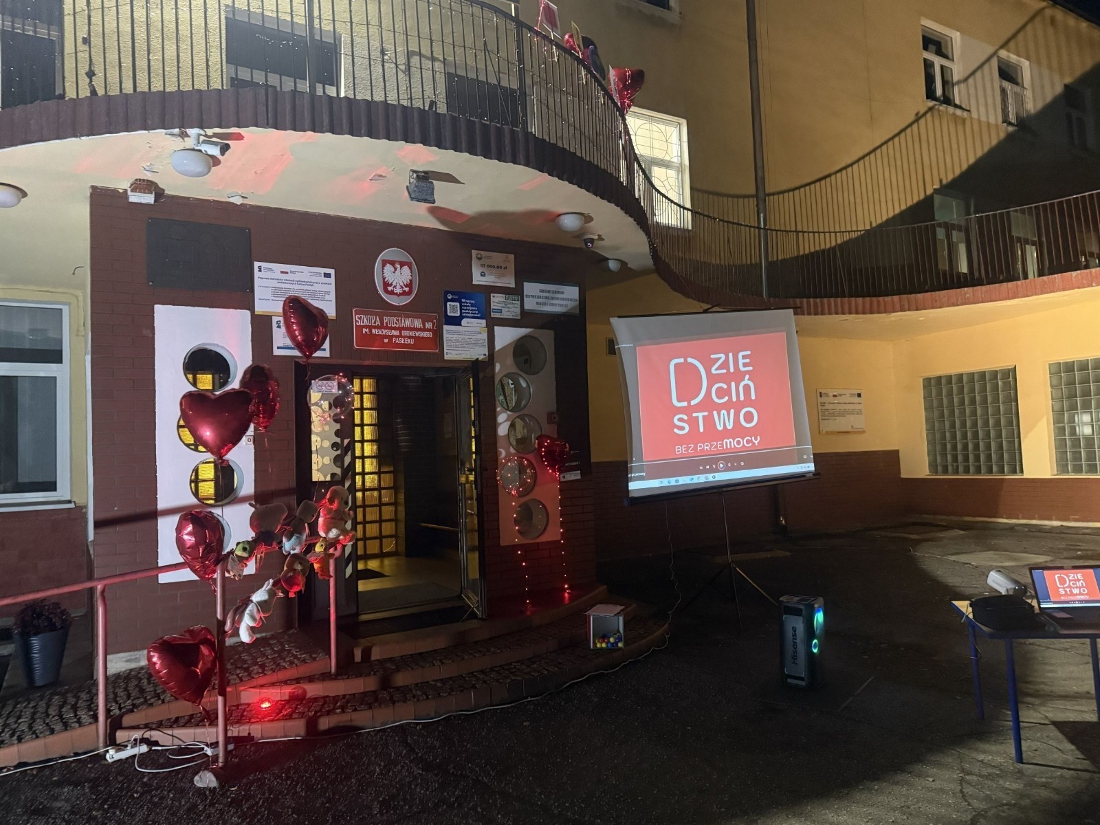 O godzinie 17:00 pod szkołą rozpoczęła się impreza, która zgromadziła około 200 osób. Szkoła została podświetlona na czerwono, co symbolizowało naszą jedność w walce z przemocą. Był to akcent podsumowujący nasze wcześ iej sze  działania w ramach Kampanii Ogólnopolskiej „Dzieciństwo bez Przemocy”. Od października uczniowie uczestniczyli w lekcjach online przygotowanych przez Fundację Dajemy Dzieciom Siłę. Rodzice również zostali objęci wsparciem w postaci webinaru online ww. fudacji.
Uczestnicy mogli wziąć udział w wspólnym śpiewaniu, quizie wiedzy o przemocy oraz tworzeniu „kamieni mocy” – symboli pozytywnej energii i wsparcia. Dodatkowo, w ramach konkursów, przyznano nagrody ufundowane przez Urząd Miejski w Pasłęku.
Ten cudowny czas umilił nam smaczny poczęstunek, w tym grochówka, pizza i ciasta, gorąca kawa i herbatka przygotowany przez panie kucharki i rodziców.
W wydarzeniu uczestniczyli również zaproszeni goście, w tym Burmistrz Pasłęka – pani Elżbieta Wasiuk,  podinspektor KP Pasłęk – pani Agnieszka Bielewicz - Piechowiak oraz przedstawiciel Kuratorium Oświaty w Elblągu – pani Teresa Bednarz, którzy wspierają nasze działania. Ich obecność była dowodem na to, jak ważna jest współpraca lokalnej społeczności w walce z przemocą wobec dzieci. Dziękujemy wszystkim, którzy wzięli udział w tym wyjątkowym dniu oraz przyczynili się do jego organizacji. Razem możemy stworzyć bezpieczne i przyjazne środowisko dla naszych dzieci!
